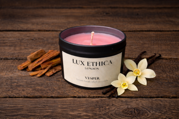 Pink candle in a black container surrounded by cinnamon sticks and flowers on a wooden surface.